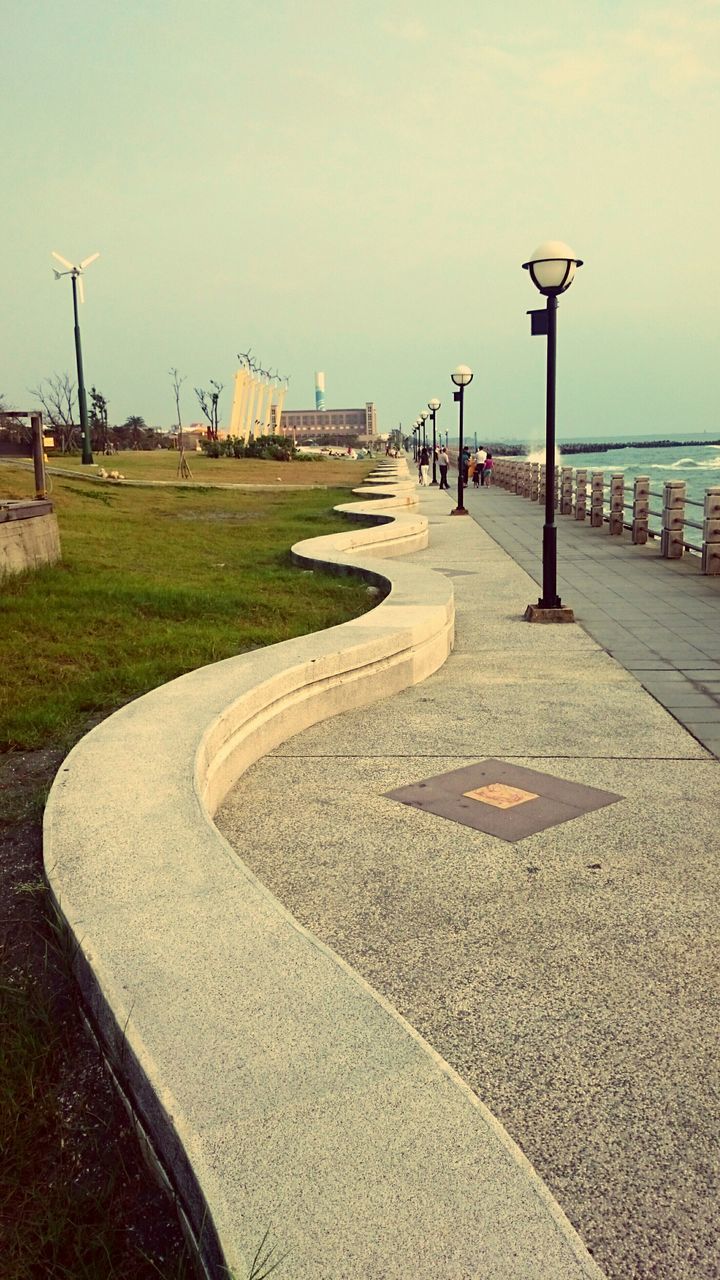 street light, clear sky, road, beach, street, sea, sky, incidental people, sunlight, water, built structure, sand, asphalt, footpath, day, outdoors, shadow, architecture, empty, road marking