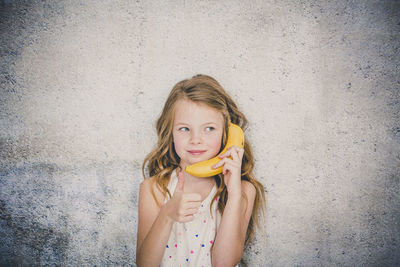 Close-up of cute girl using banana as phone while playing against textured wall