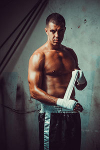 Portrait of shirtless male athlete wearing hand wrap while standing in darkroom