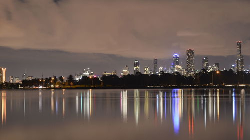 Albert park lake by illuminated buildings against cloudy sky at night