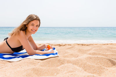 Portrait of smiling woman holding sunscreen bottle while lying on towel at beach