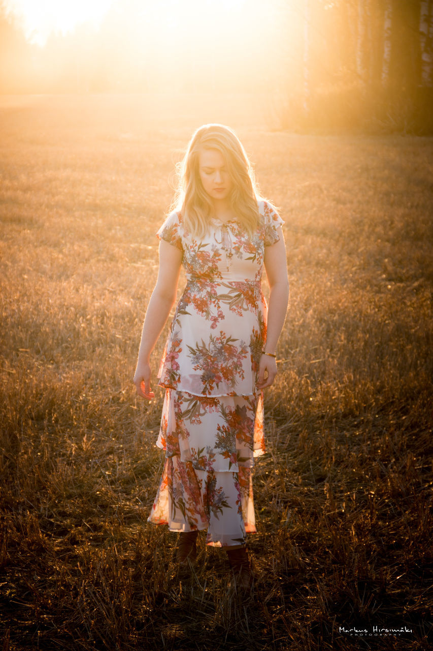 field, one person, leisure activity, land, sunlight, plant, lifestyles, real people, sunset, nature, women, grass, standing, three quarter length, front view, sky, casual clothing, young adult, full length, hairstyle, fashion, lens flare, beautiful woman, outdoors