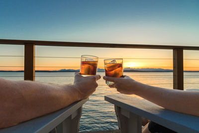 Cropped hands of friends holding whiskey in glass on bench by sea during sunset against sky
