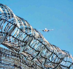 Low angle view of plane landing against clear blue sky with barbed wire fence 