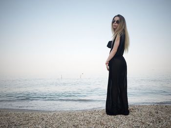 Full length of young woman wearing black dress while standing on shore at beach against sky