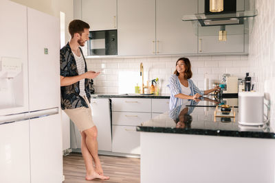 Smiling woman making coffee while looking at man using phone in kitchen