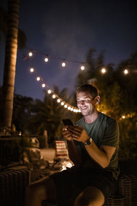 Smiling man texting while sitting outdoors during sunset