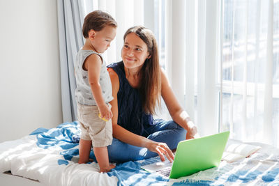Happy mother working from home online on laptop with toddler kid together