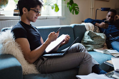 Young man studying while friend using social media on sofa at home