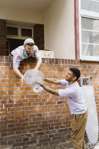 Smiling gay couple helping each other while holding electric fan over brick wall