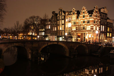 Bridge over river by illuminated buildings in city at night