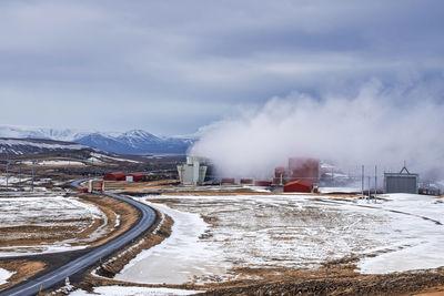 Steaming cooling tower at krafla geothermal power plant by road against sky