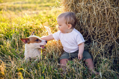 Gay boy kid blonde in white tank top sitting on a field of hay next to stack with  dog during sunset