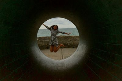 Woman jumping at beach seen through pipe