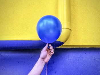 Close-up of hand holding blue balloon against yellow and purple wall.