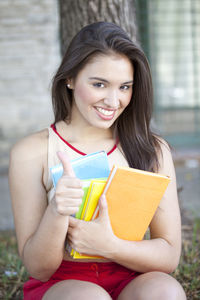 Portrait of smiling young woman gesturing thumbs up while holding books