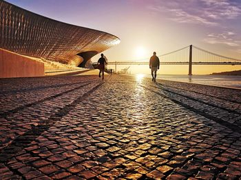 People walking on bridge in city against sky during sunset