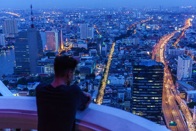 Rear view of man standing in balcony by illuminated cityscape at dusk