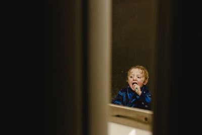 Boy brushing teeth reflecting on mirror seen through doorway