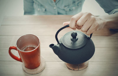 Black woman wearing a denim shirt serving tea. wooden table. red cup, black teapot.