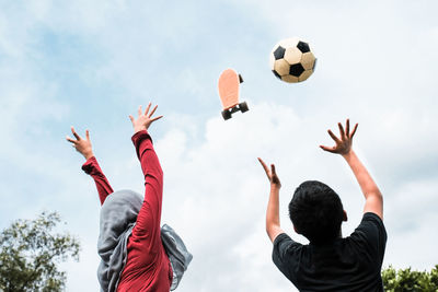 Rear view of siblings with arms raised against skateboard and soccer ball in mid-air