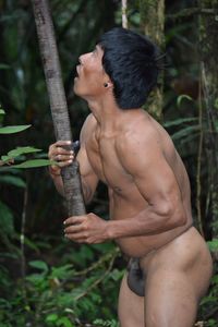 Midsection of shirtless man holding tree in forest