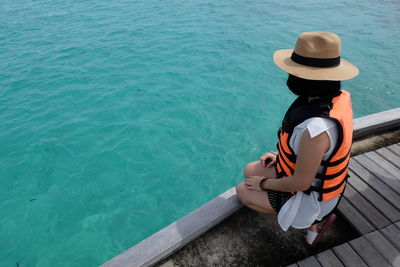 High angle view of woman wearing life jacket and hat while sitting on pier by sea