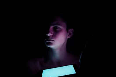 Close-up of teenage boy with illuminated mobile phone looking away in darkroom