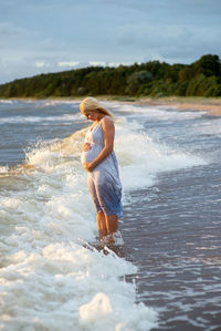 Sea waves rushing towards pregnant woman touching abdomen while standing at shore