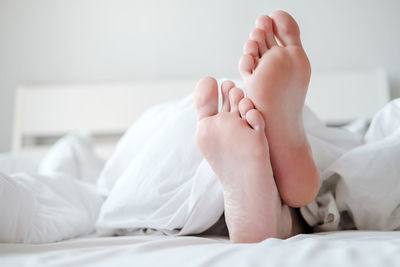 Men's feet under white blanket, in bed, in comfortable apartment in natural light, with copy space.