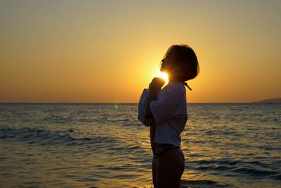 Side view of woman praying while standing on beach against sky during sunset