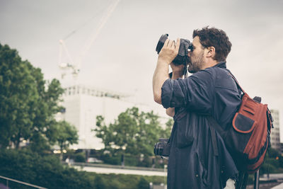 Side view of man with backpack photographing while standing against sky