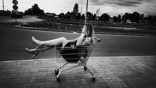 Side view of woman sitting in shopping cart on footpath
