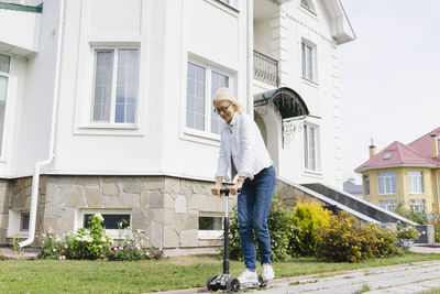 Happy senior woman riding push scooter in front of house