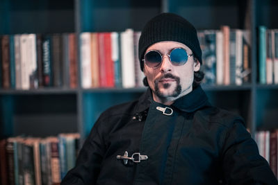 Portrait of handsome man wearing sunglasses while sitting against bookshelf