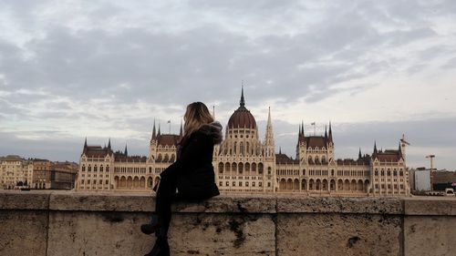Woman sitting on retaining wall against hungarian parliament building