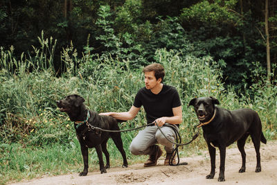 Millennial man sitting with his two black dogs on alert in the forest
