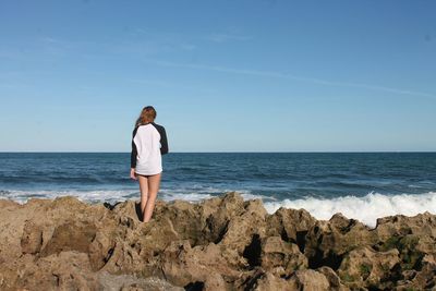 Rear view of woman standing on rocky sea shore against sky during sunny day