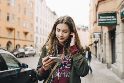 Smiling girl listening music through smart phone while walking on sidewalk in city