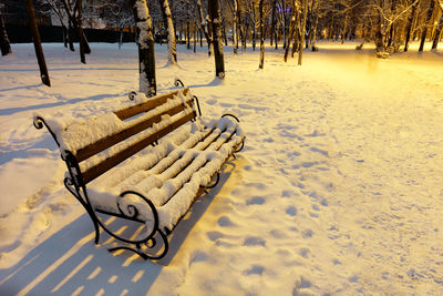 Empty brown wooden bench with metal decorative railing in the snow on winter evening in a city park.