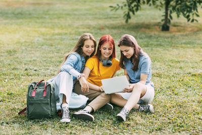 Teenage girls are sitting on a green lawn in a park with a backpack and a digital tablet 