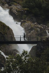 Side view of man walking on footbridge against waterfall