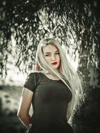 Portrait of young woman with long hair standing against tree in park