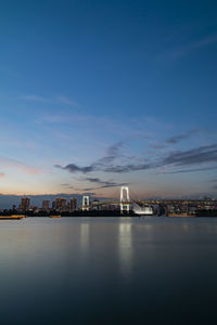 Illuminated city by sea against sky during sunset