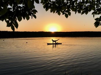 Silhouette women doing yoga over raft on lake against sky during sunset