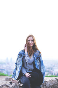 Portrait of young woman sitting on retaining wall against sky