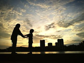 Silhouette mother and son holding hands while standing in city against sky during sunset