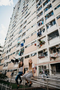 Full length of smiling female friends sitting on railing against building in city