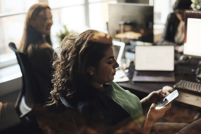 Side view of female entrepreneur text messaging through smart phone seen through glass at office