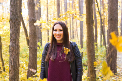 Smiling young woman standing by tree trunk in forest during autumn